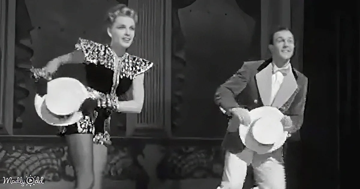 A woman in a patterned dress and a man in a suit are performing a dance routine, both holding white hats.