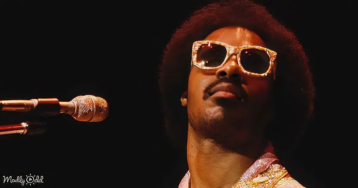 A man with an afro and sunglasses stands in front of a microphone, wearing a glittery outfit.