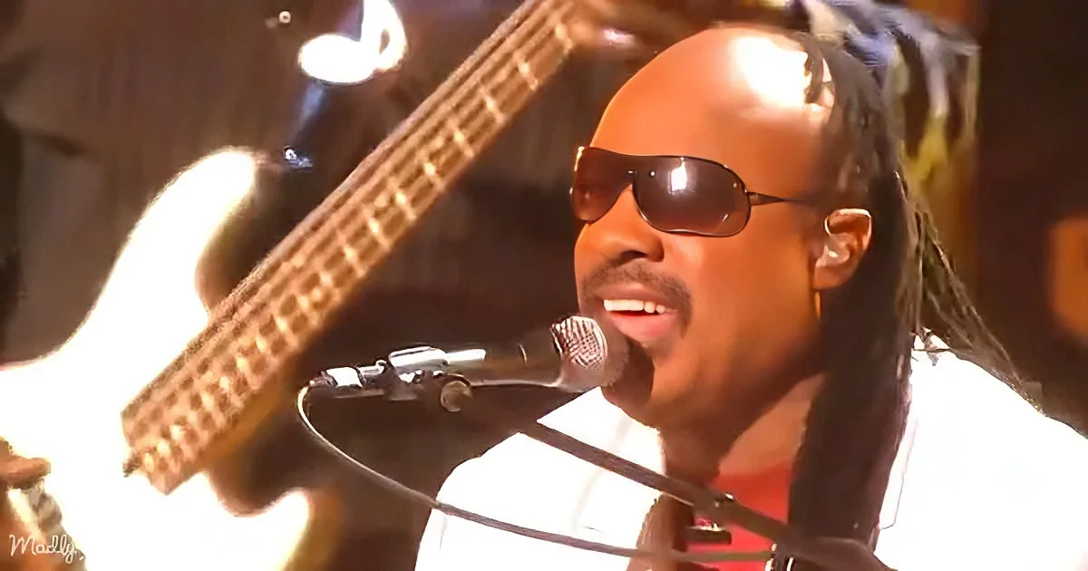 A man with sunglasses and dreadlocks sings into a microphone, with a guitar in the background.