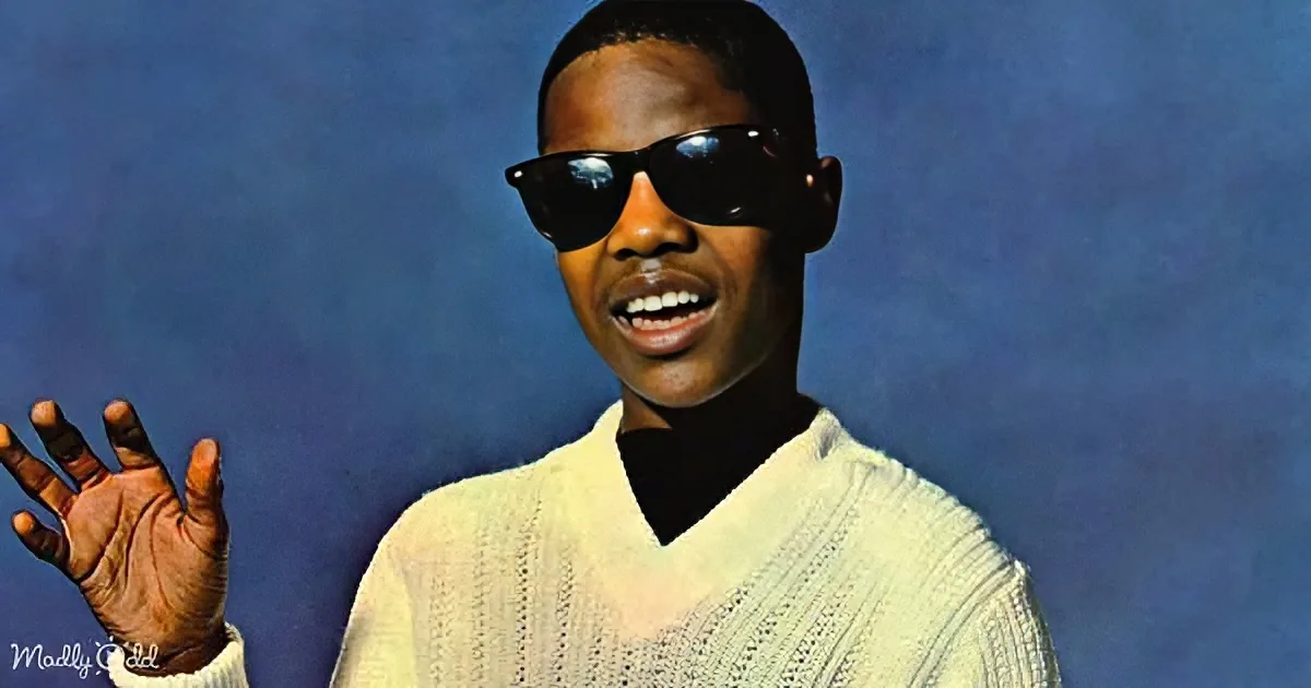A person wearing sunglasses and a white sweater gestures with their hand against a blue background.