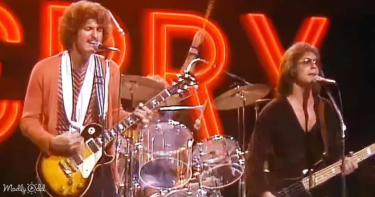 A guitarist in a brown shirt plays a yellow guitar on stage with a drummer and another guitarist in a black outfit.
