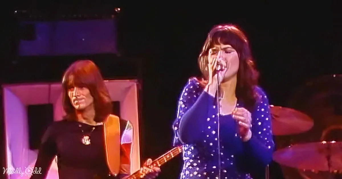 A woman in a blue sequined dress sings into a microphone while another plays guitar on stage.