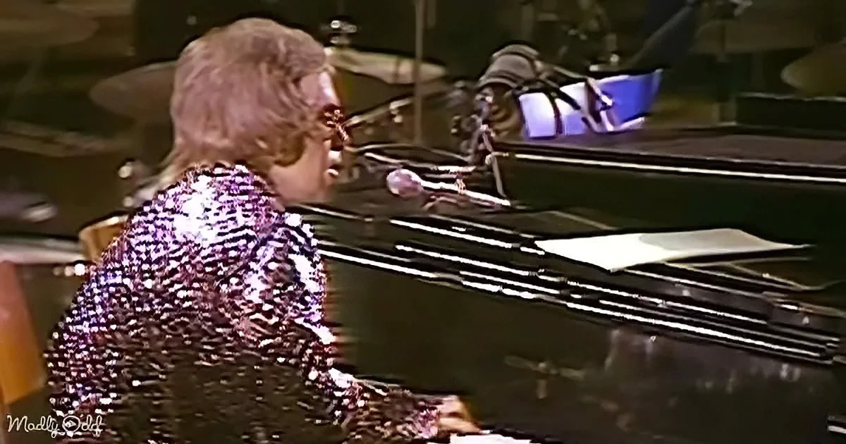 A person in a glittery outfit plays piano and sings into a microphone on stage.