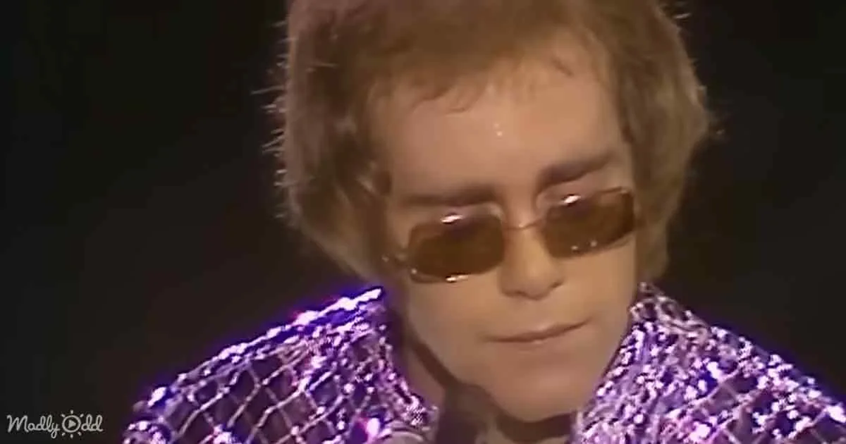 A person wearing sunglasses and a sparkly purple outfit is seen against a dark background.