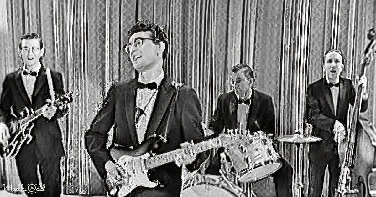 A man in a suit plays guitar while a band in tuxedos performs behind him.