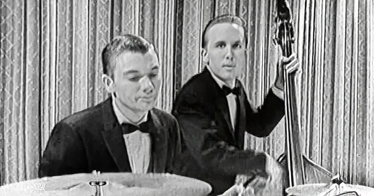 A man in a tuxedo plays drums while another in a suit holds a bass, both in front of a curtain backdrop.