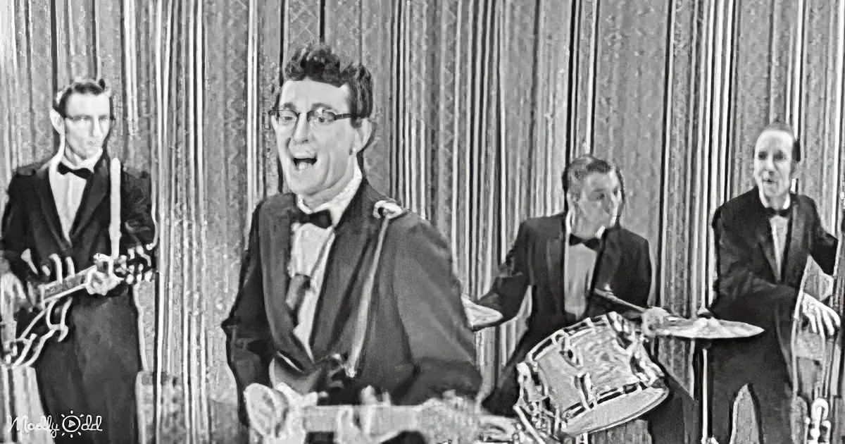 A man in a tuxedo plays guitar while a band, including a drummer and guitarist, performs.