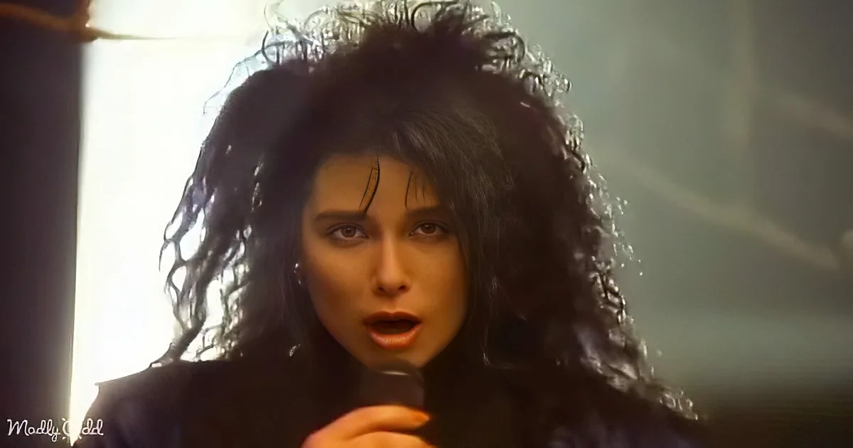 A person with long, dark, curly hair sings into a microphone, illuminated by a bright light.