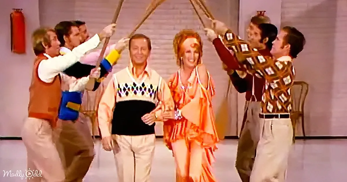 A man in a colorful sweater is surrounded by dancers in vibrant costumes, holding sticks above his head.