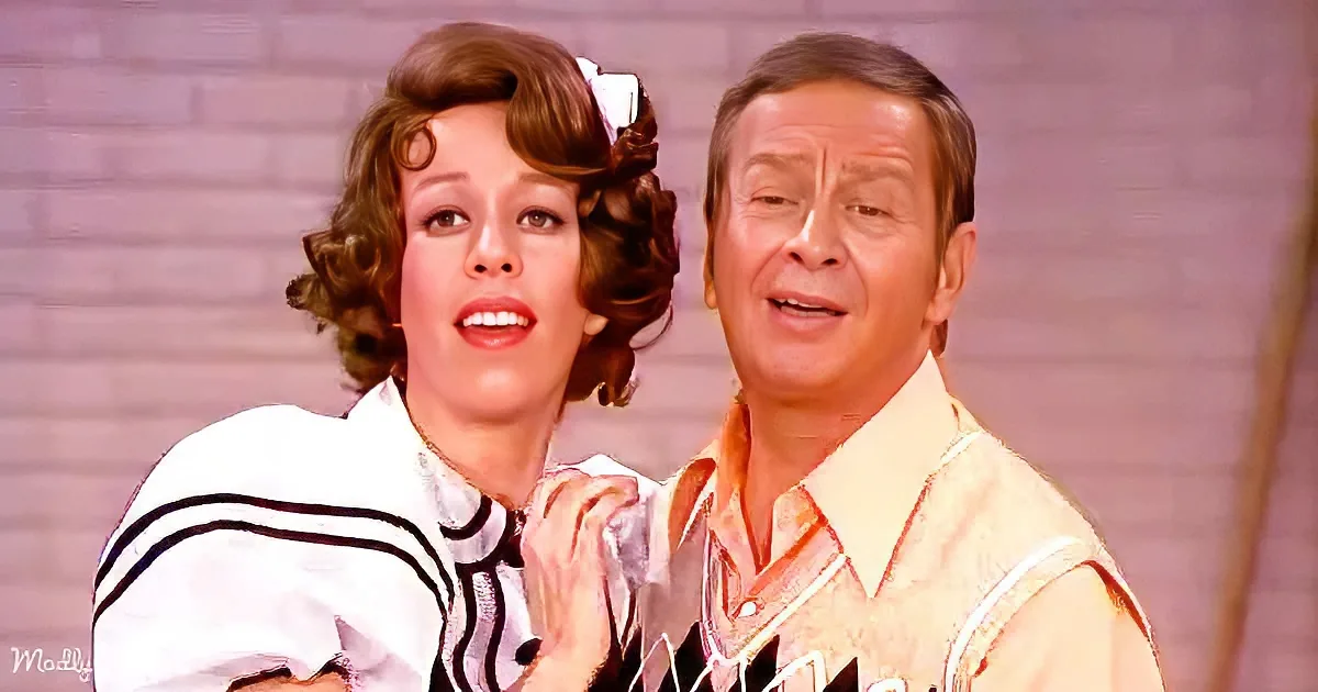 A man and woman in colorful costumes pose together, smiling, against a brick wall backdrop.