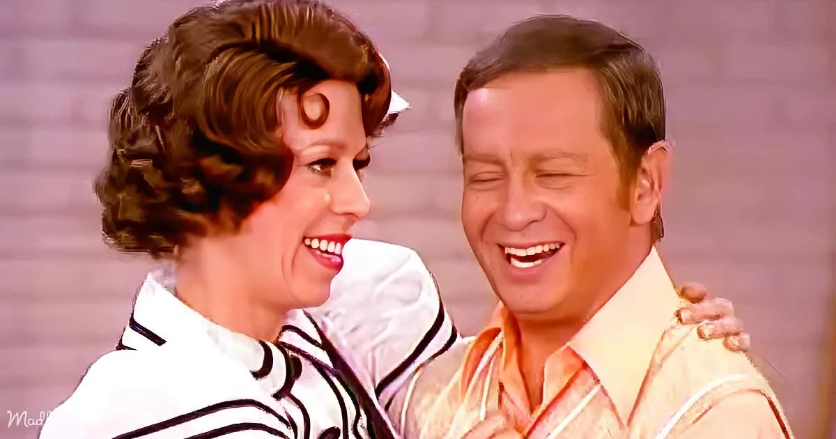 A man and woman are embracing, both smiling, with the woman's arm around the man's shoulder.