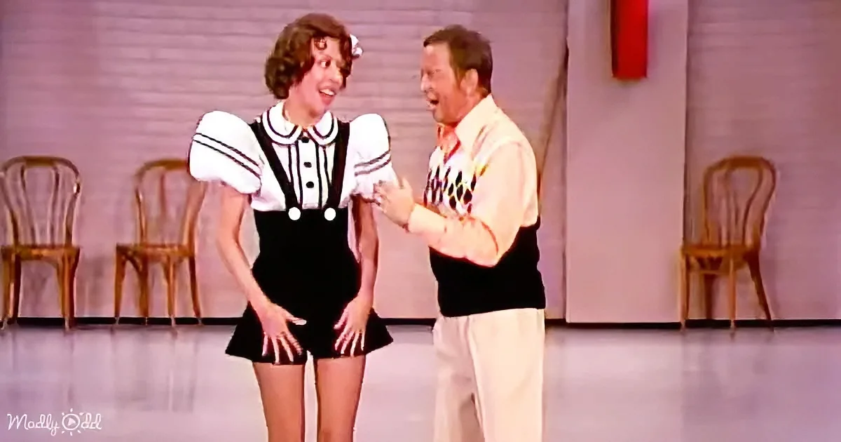 A woman in a sailor-style dress and a man in a vest and white pants are interacting on stage.