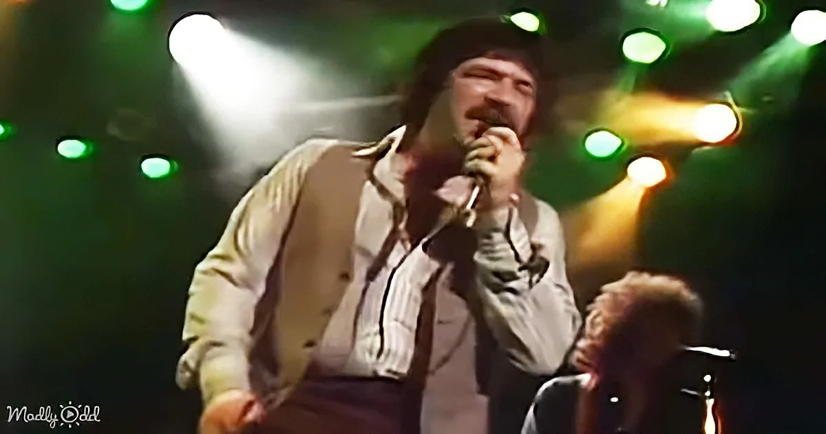 A man with a beard and mustache sings into a microphone on stage with colorful lights.