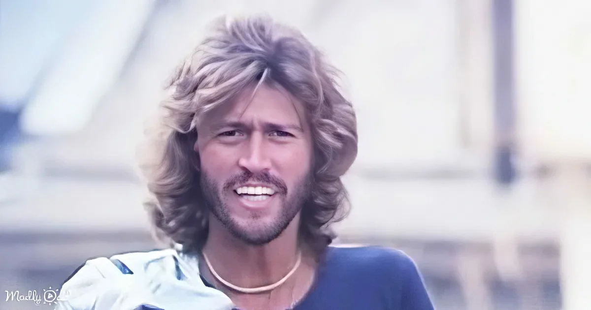 A man with long, wavy hair and a beard, wearing a white shirt and a gold necklace, smiles in front of a blurred background.