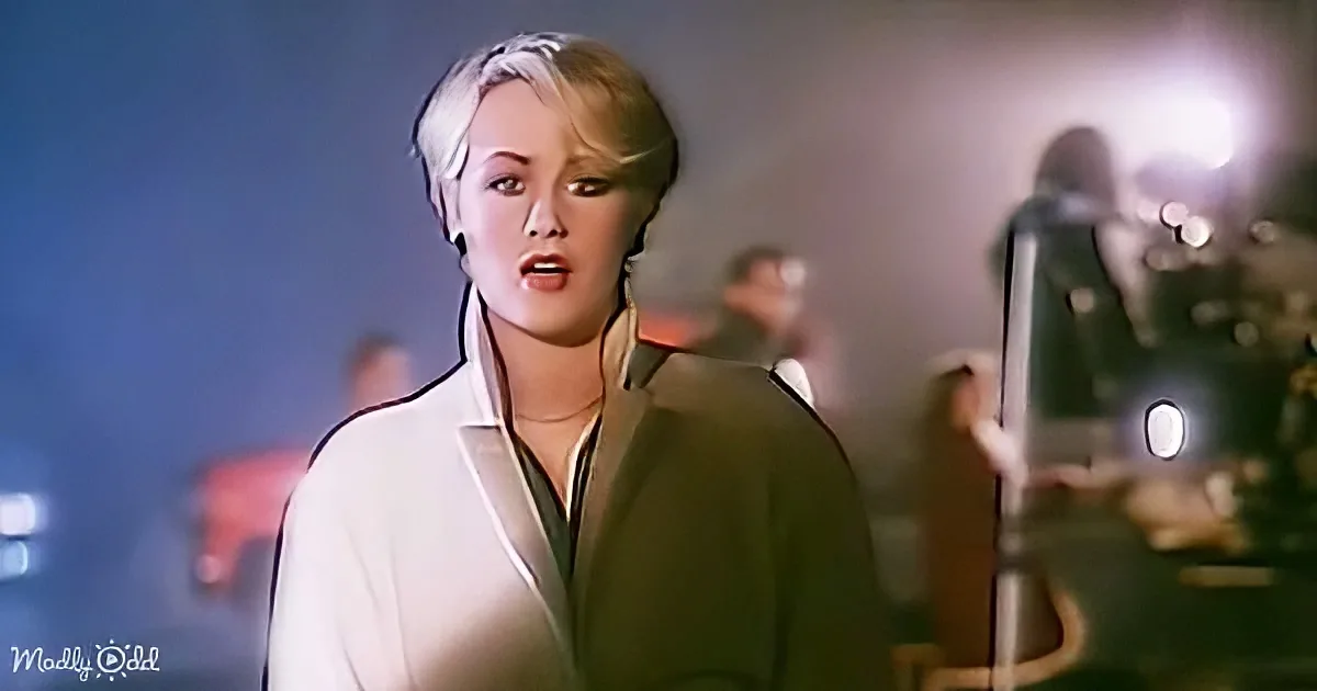 A woman in a white blazer and green shirt stands in a dimly lit room, holding a phone.
