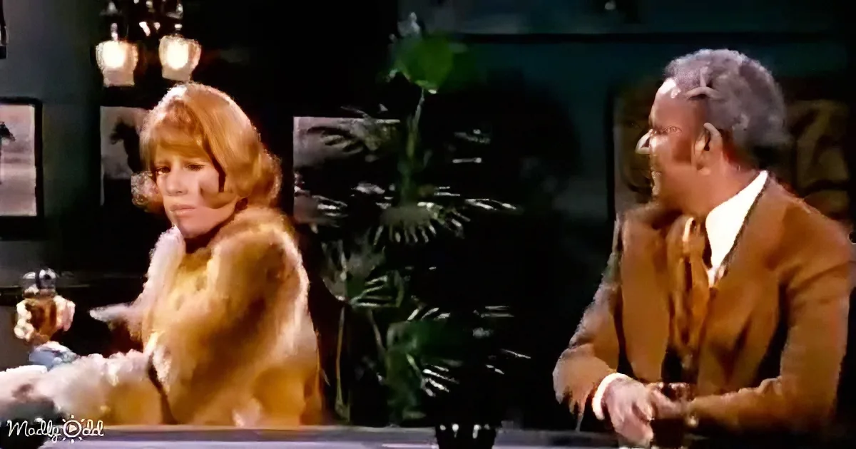 A woman in a fur coat sits at a table, gesturing, while a man in a suit looks on, in a dimly lit room with plants.