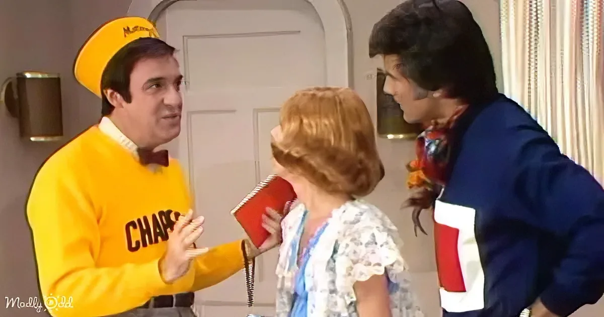 A man in a yellow "Chap" uniform gestures while holding a red notebook, interacting with another man and a woman.