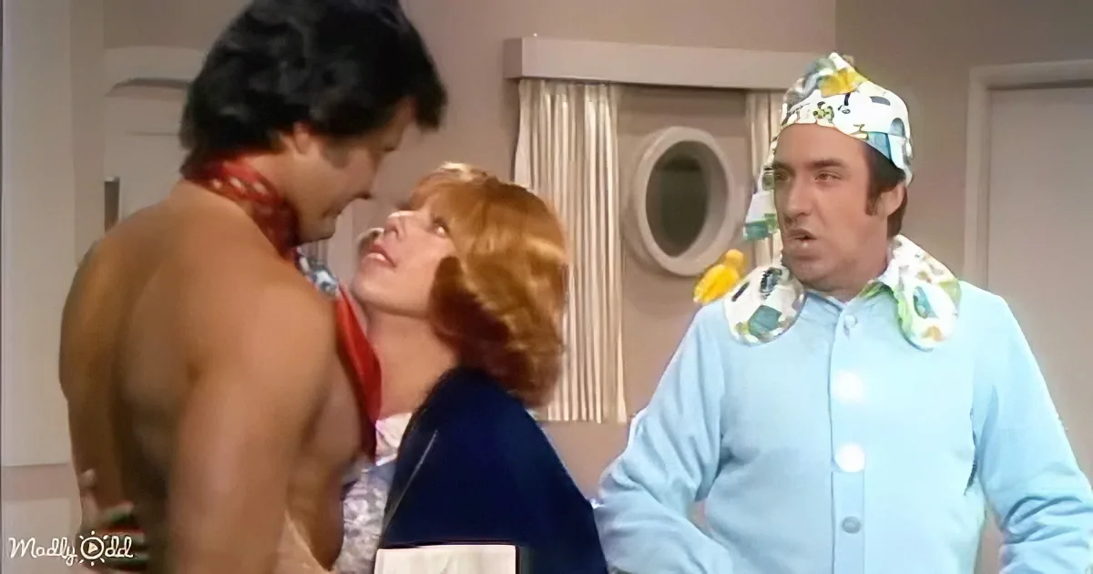 A shirtless man leans toward a woman, while another man in a floral shirt looks on, in a room with a window and curtain.