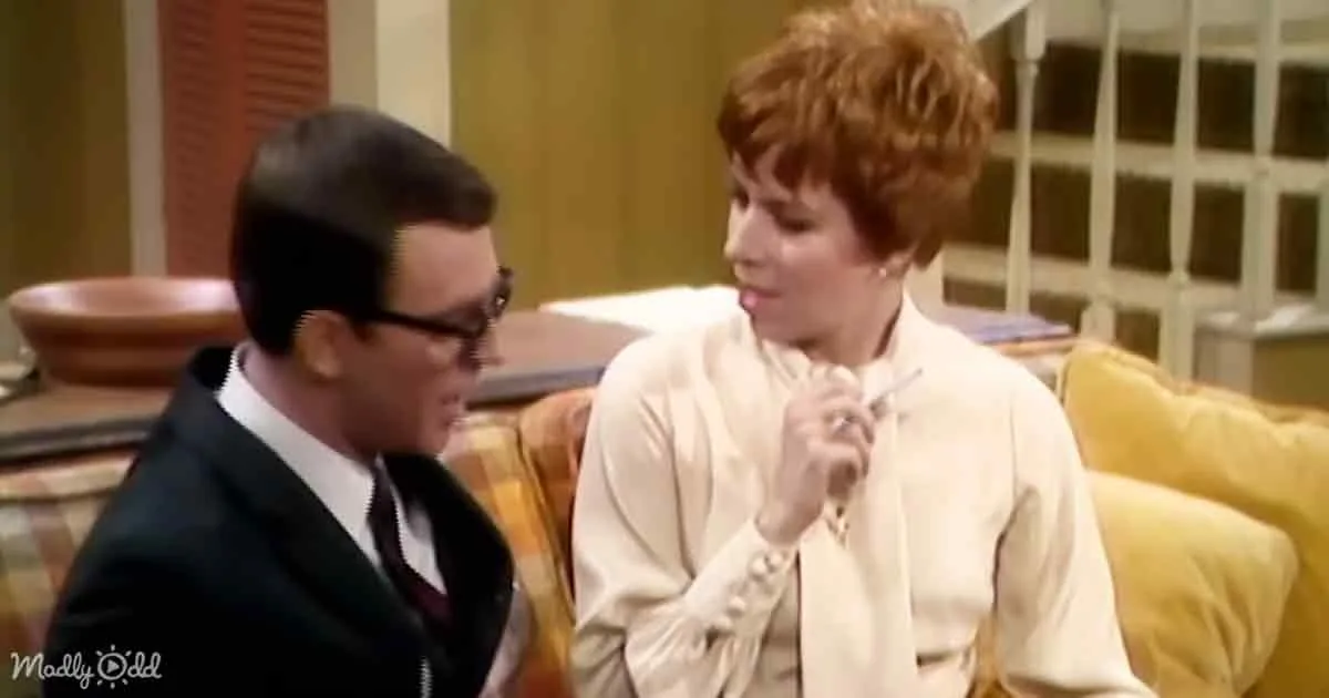 Woman in white blouse holding pen, looking at man in suit with glasses in a room with yellow couch.