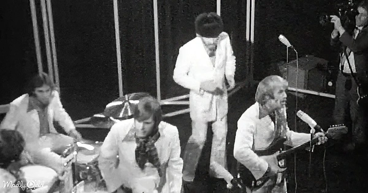 A man in a white suit sings on stage, surrounded by band members and microphones.