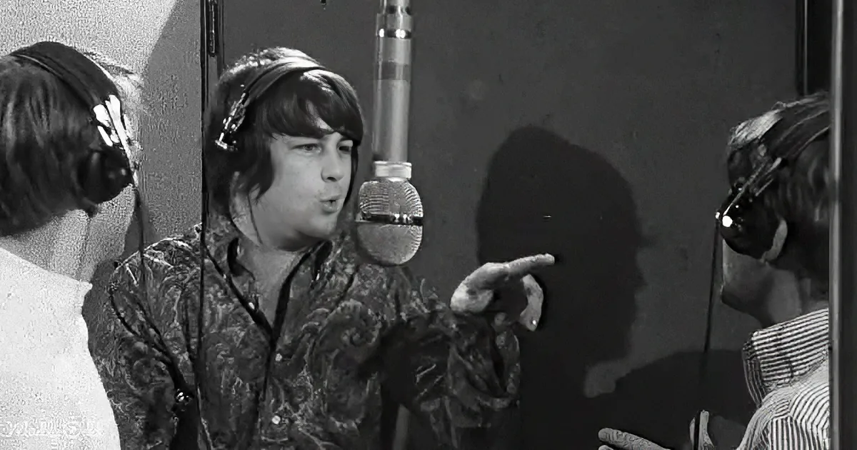 A person in a patterned shirt sings into a microphone, wearing headphones, with another person in the background.