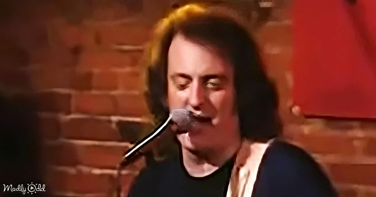 A person with long hair sings into a microphone, wearing a black shirt and white vest, against a brick wall backdrop.