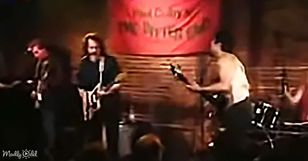 A shirtless man plays guitar on stage, surrounded by other musicians and a large red sign.