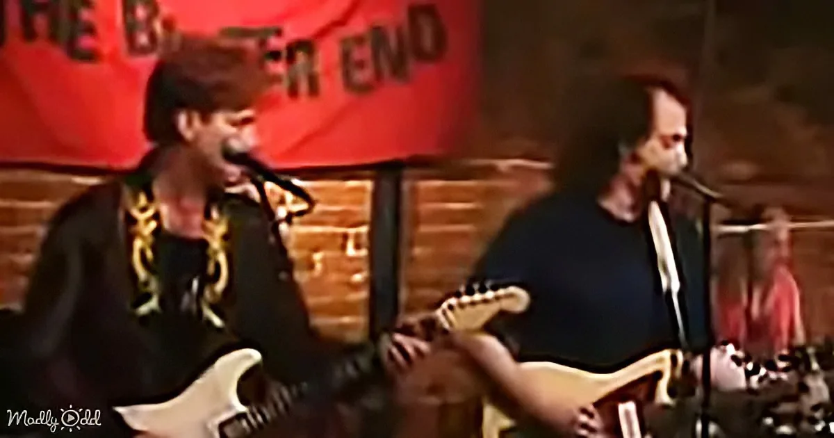 Two musicians play guitars on stage, one singing into a microphone, with a red banner in the background.