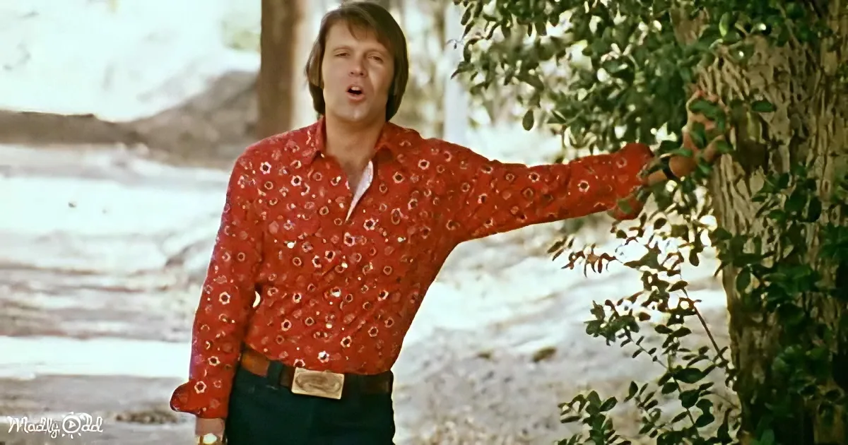 A man in a red floral shirt and jeans stands outdoors, pointing to a tree.