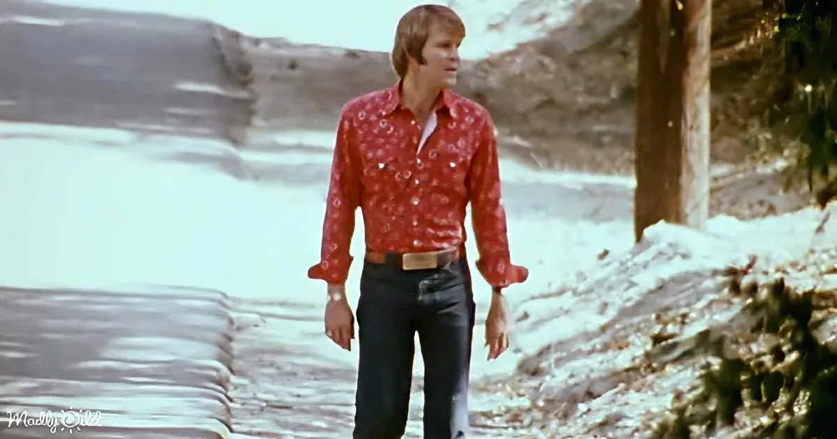 A man in a red patterned shirt and jeans walks along a snowy path near a body of water.