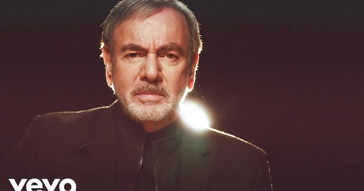 A man with short gray hair and a beard, wearing a black jacket, stands against a dark background with a bright light...