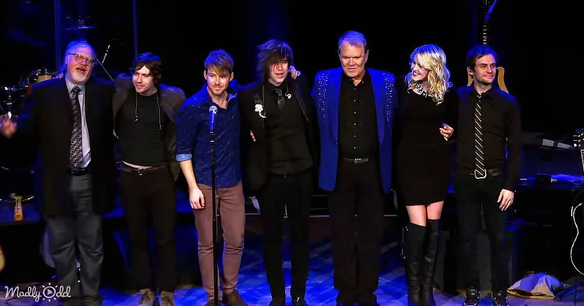A group of seven people, including a man with a microphone, stand on stage in formal attire, smiling and posing together.