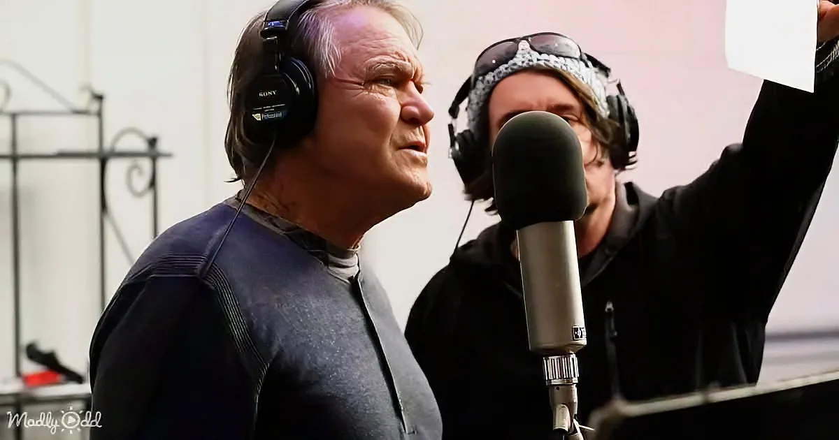 An older man wearing headphones sings into a microphone while another person holds a piece of paper.