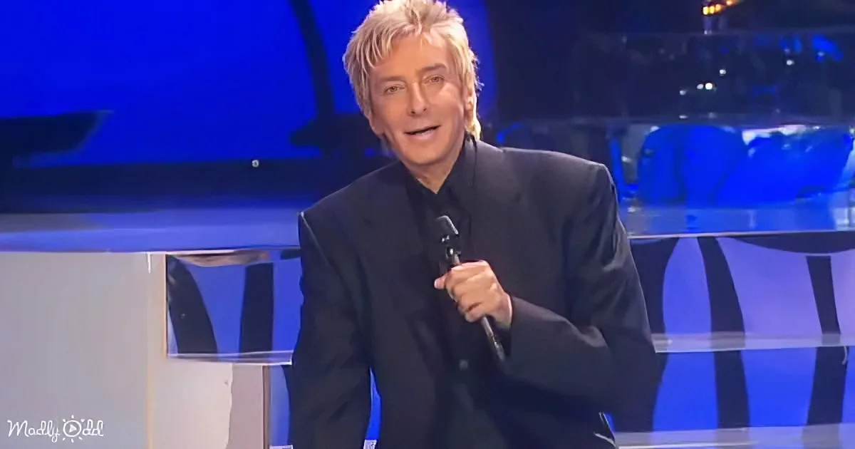 A man in a black suit holds a microphone on stage with blue lighting.