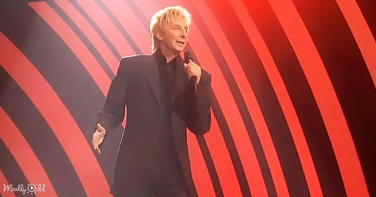 A person in a black suit holds a microphone, standing against a red and black striped background.