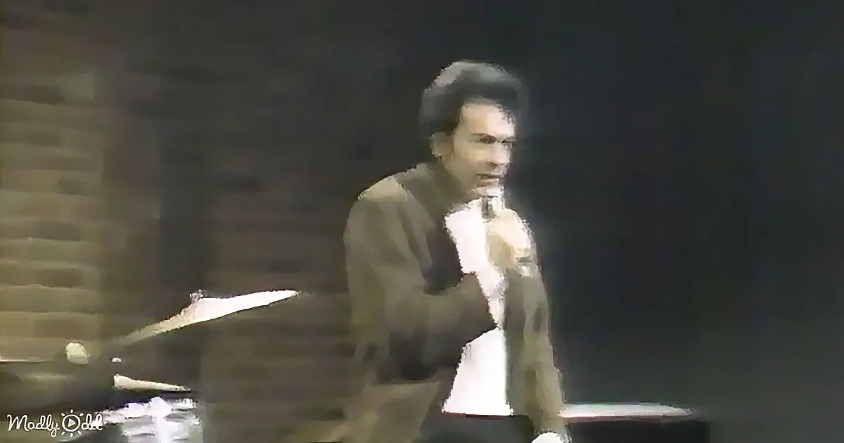 A man in a suit holds a microphone, standing in front of a dark background.