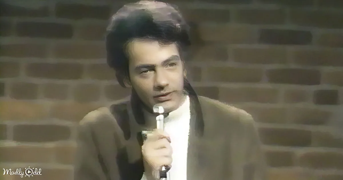 A man in a suit holds a microphone, standing against a brick wall.