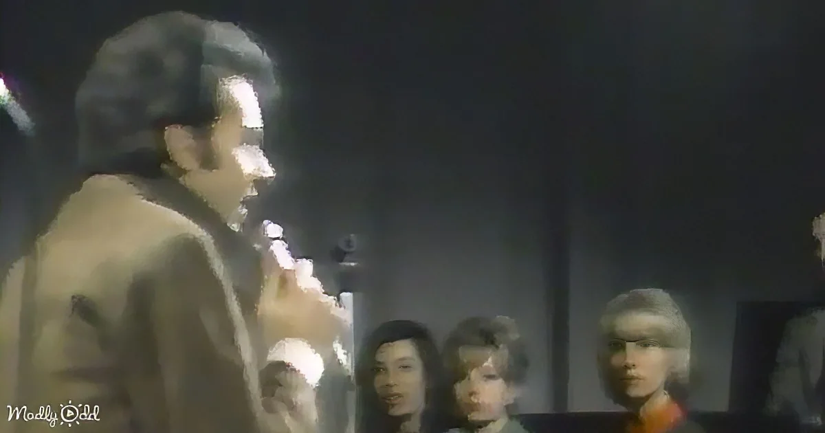 A man in a suit speaks into a microphone, with a blurred audience in the background.
