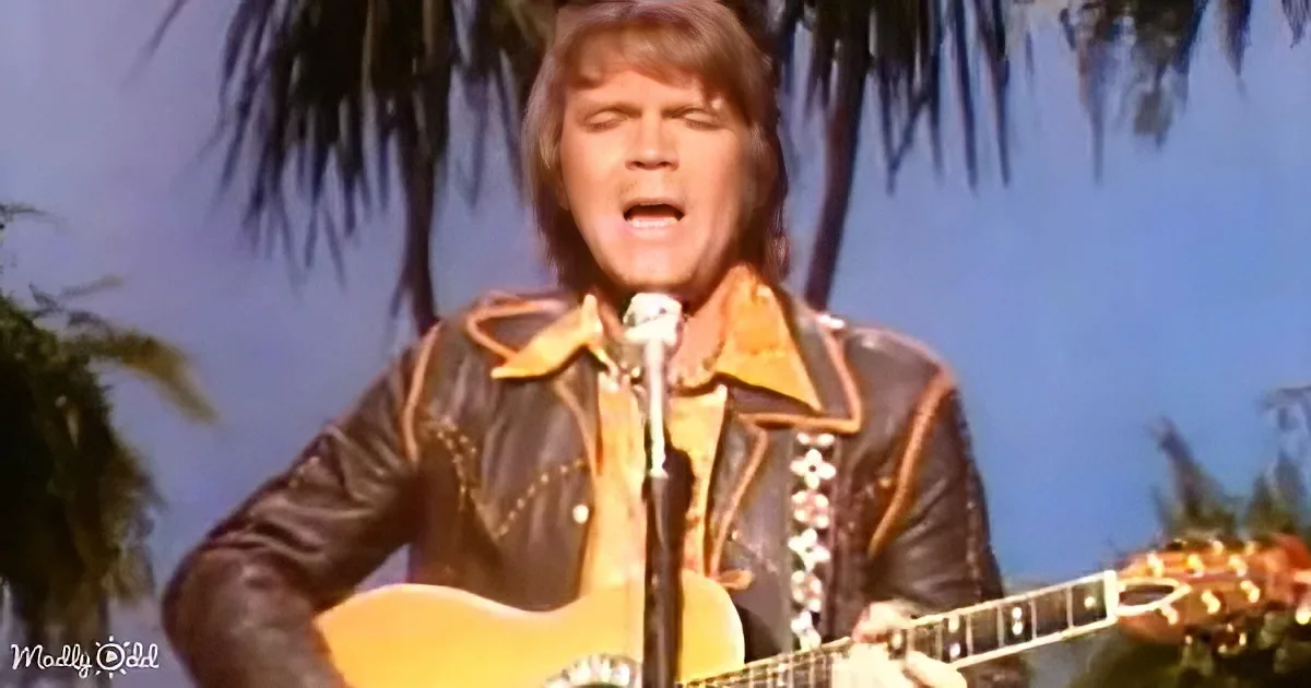 A man in a leather jacket plays an acoustic guitar on stage with palm trees in the background.
