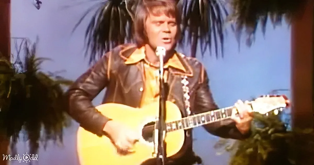 A person in a leather jacket plays an acoustic guitar on stage with a microphone and palm trees in the background.