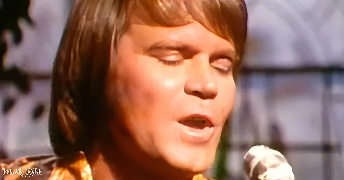A person with shoulder-length brown hair and a yellow patterned shirt is singing into a microphone.