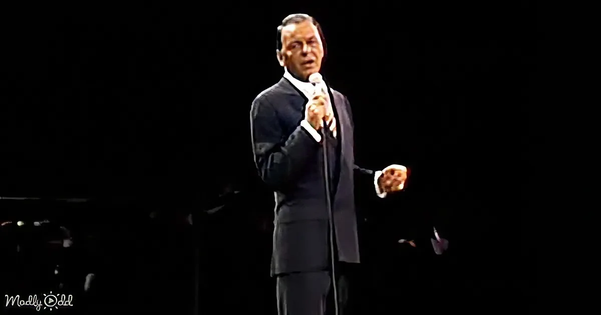 A man in a dark suit sings into a microphone on stage, with a dark background and a piano partially visible.
