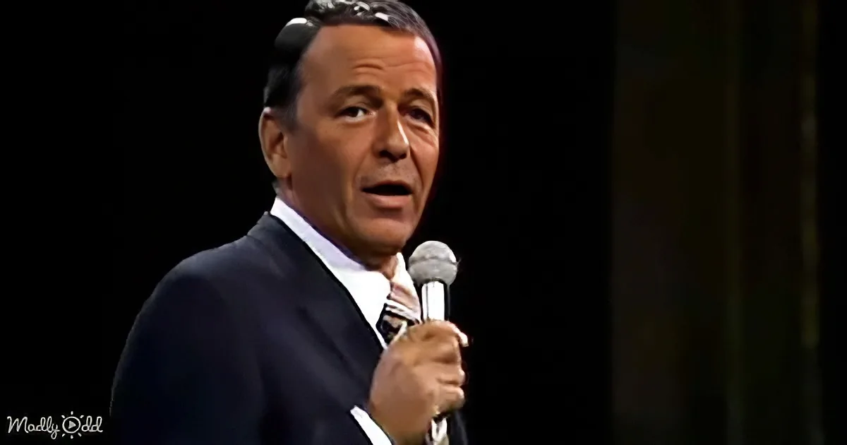 A man in a suit holds a microphone, singing on stage with a dark background.