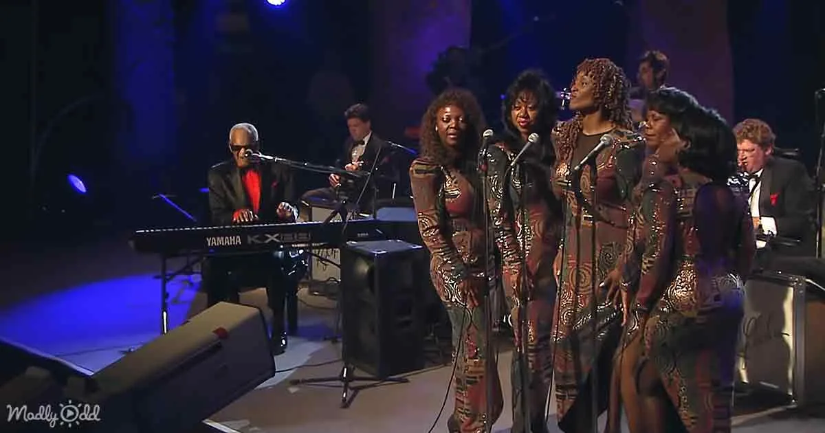 A group of women in patterned dresses sing on stage, accompanied by a pianist and other musicians.