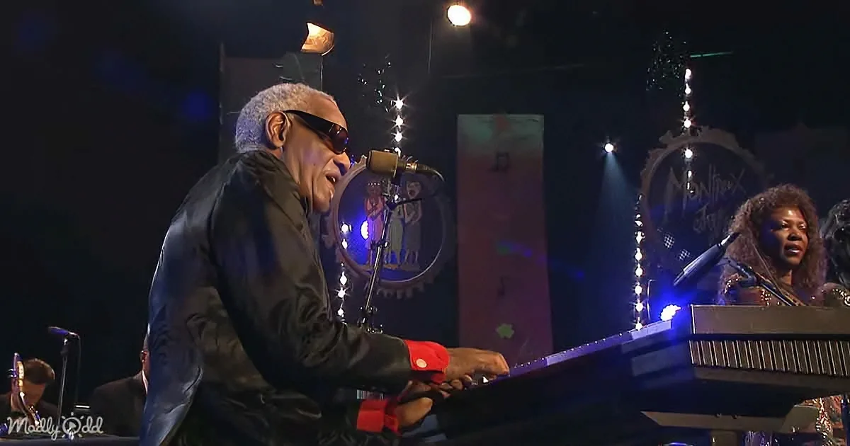 A man in sunglasses plays piano on stage, illuminated by blue lights, with a band and audience in the background.