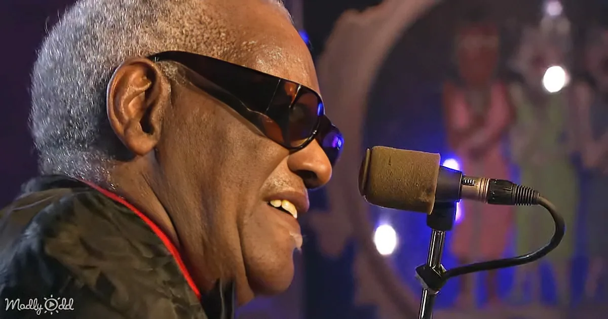 An older man with gray hair and sunglasses sings into a microphone on stage.