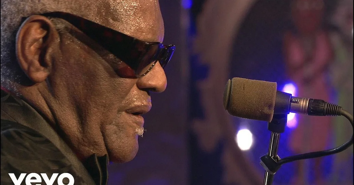An older man in sunglasses plays a keyboard on stage, smiling, with a microphone and musical backdrop.