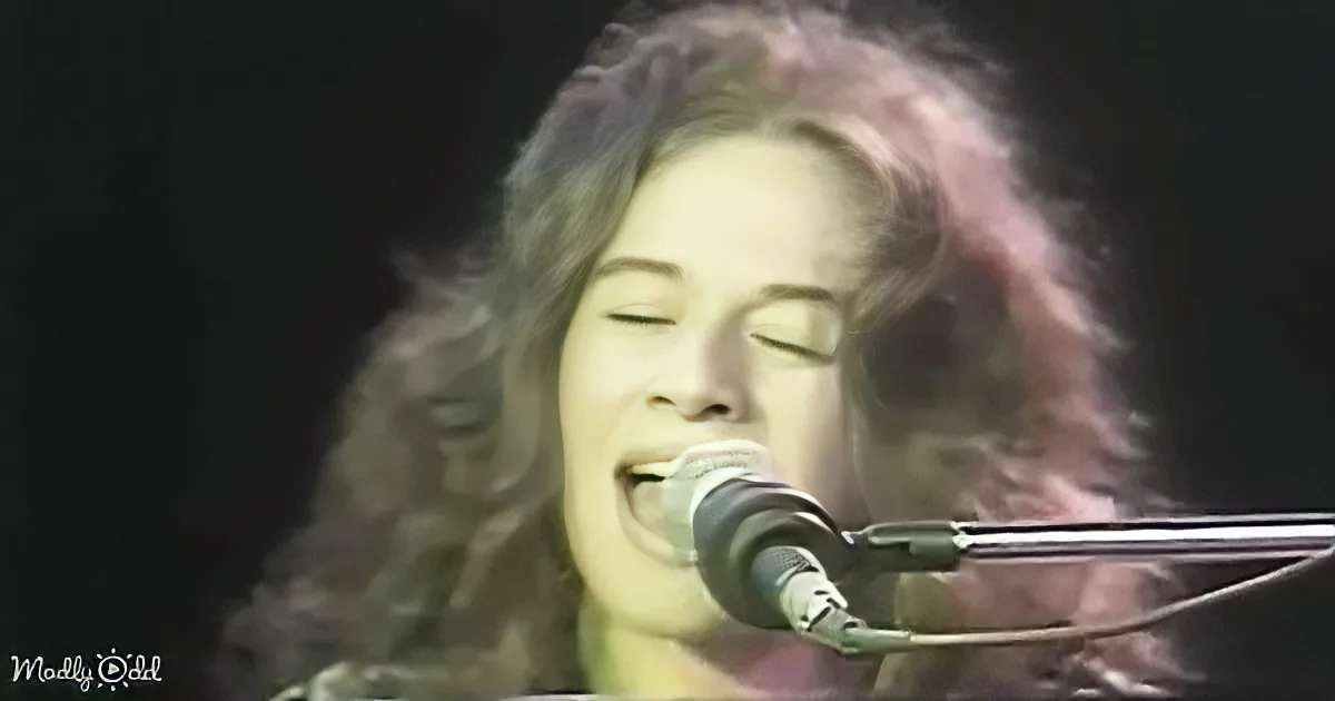 A person with long, curly hair sings into a microphone, eyes closed, in a dimly lit setting.