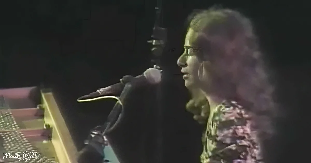 A person with long hair is singing into a microphone, wearing a patterned shirt.