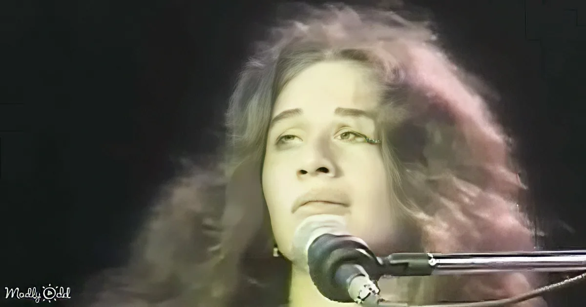 A person with long hair is singing into a microphone, their face illuminated against a dark background.